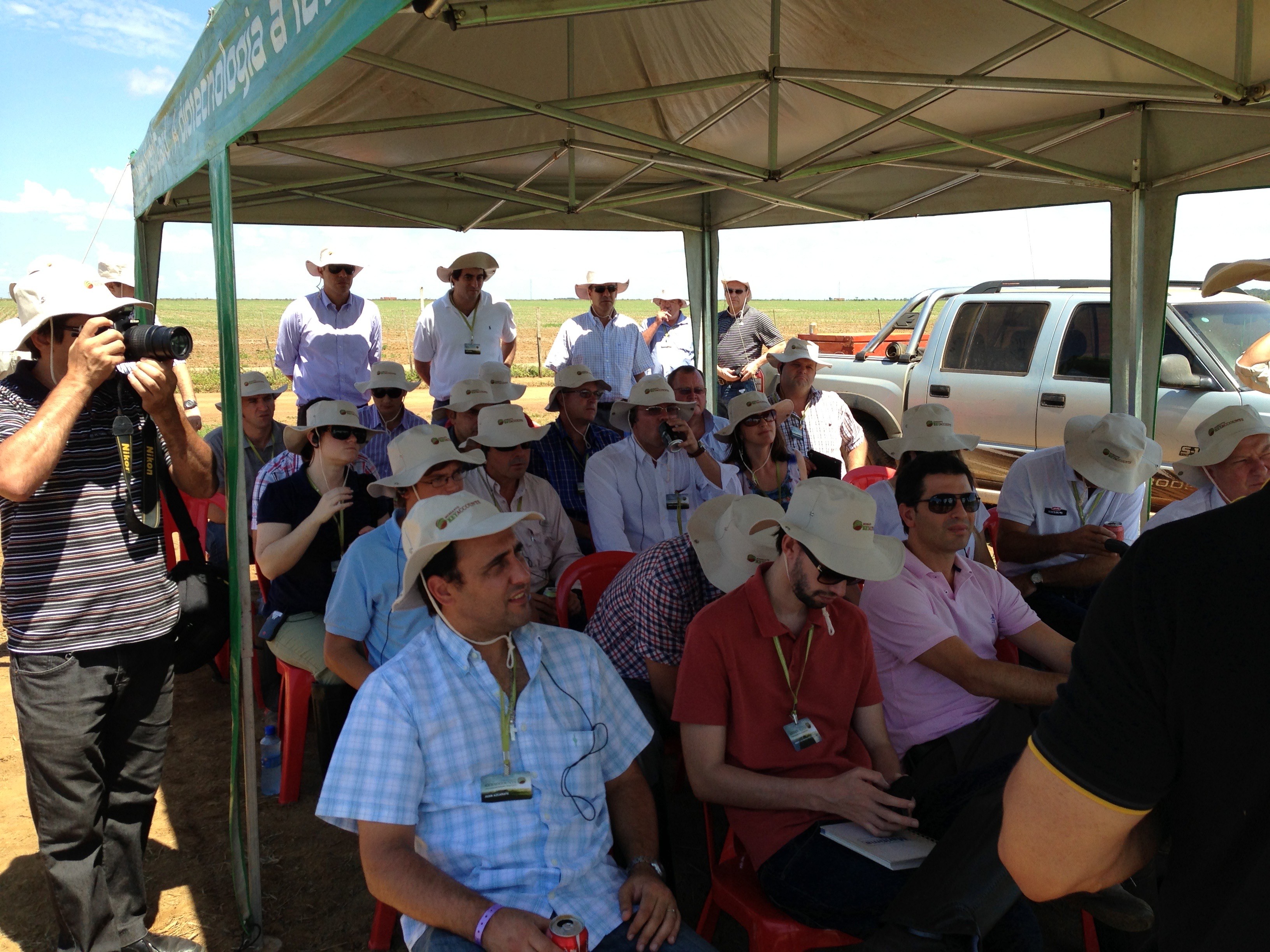 Growers and distributors seated under a branded tent at a field day, listening to a technology presentation with a crop trial behind them